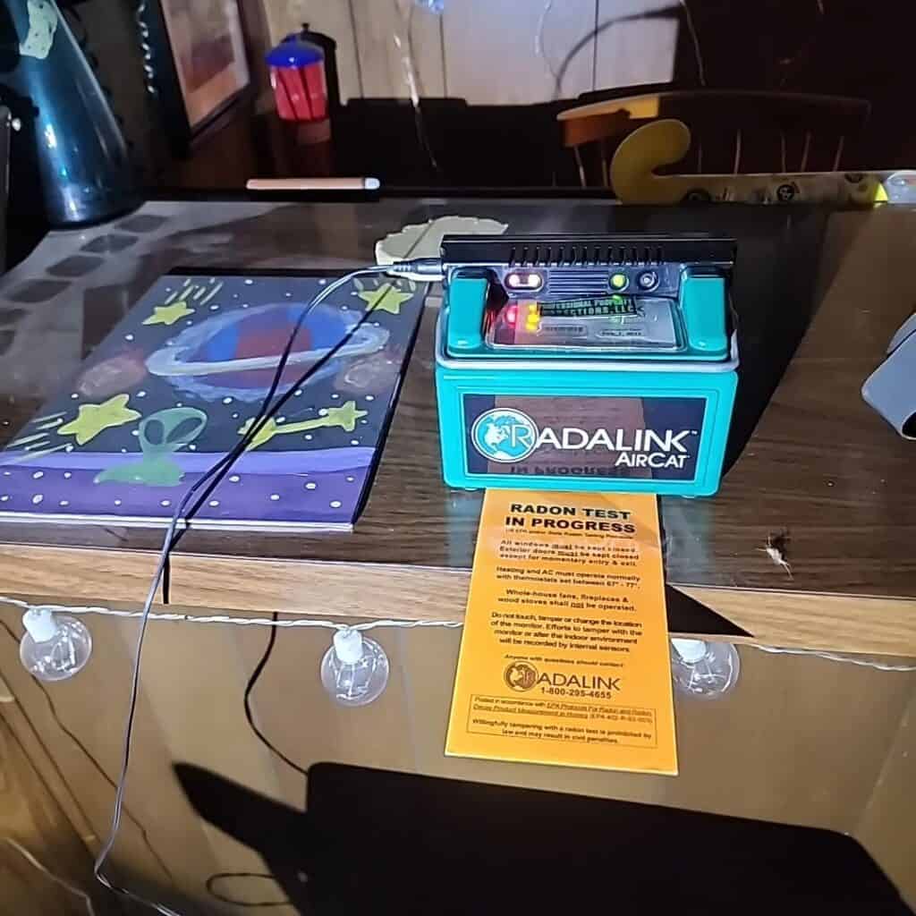 Radon testing device on a wooden table with warning notice and colorful mat.